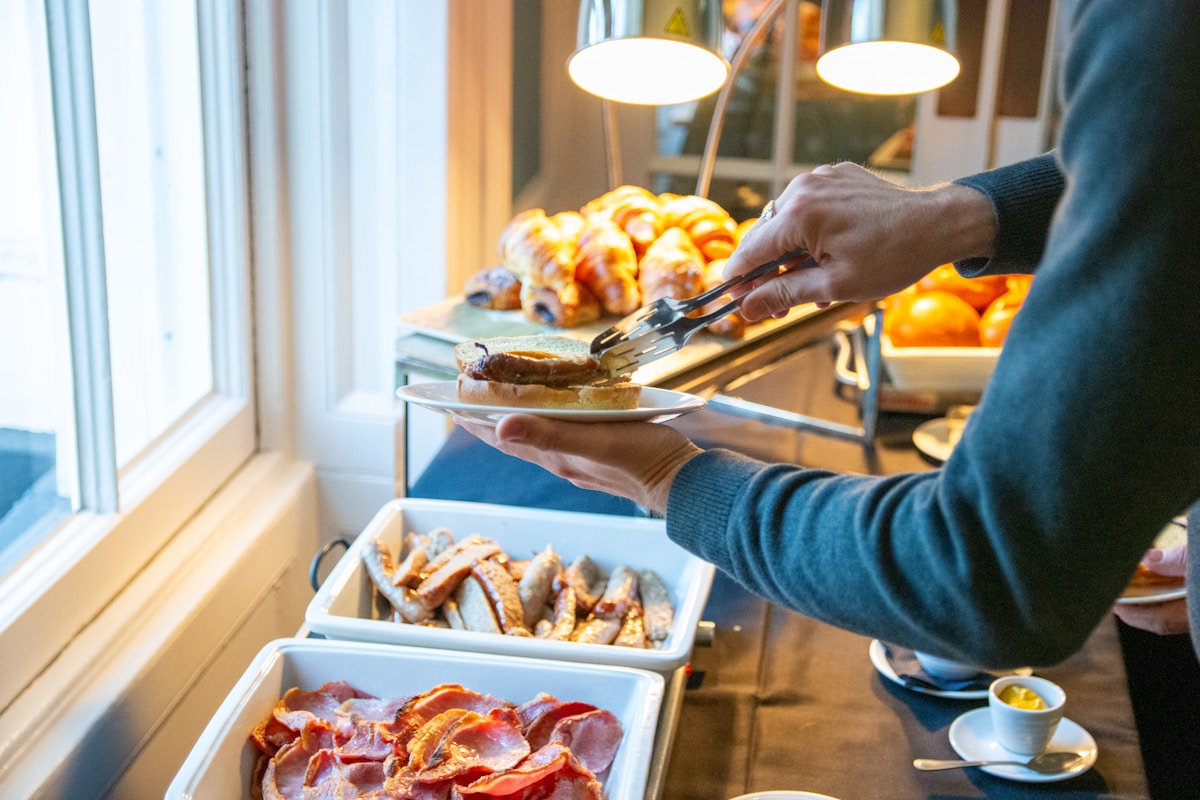A guest serving themselves from a warmly lit hotel breakfast buffet, plate in hand at the carving and pastry station