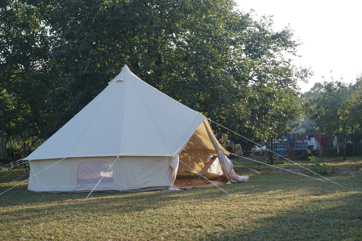 A canvas glamping tent on a grass lawn at golden hour with guests gathered at tables in the background