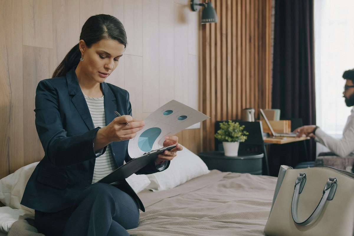 A hotel executive reviewing performance charts on a clipboard in a warm, wood-paneled guest room while a colleague works at the desk behind her